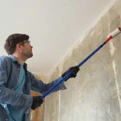 Mężczyzna w okularach ochronnych i rękawiczkach nakłada grunt na ścianę wałkiem. To pierwszy krok, jak zrobić grunt do ścian, by przygotować je do malowania.
