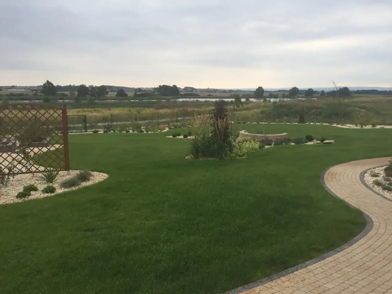 Piękna, soczysta trawa gazonowa co to za cud natury! Wokół niej kamienne ścieżki i rabaty, a w tle malowniczy krajobraz z wodą.